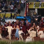 A crowded outdoor stage performance during a Super Bowl event featuring singers in white and blue outfits accompanied by a brass band in maroon suits.