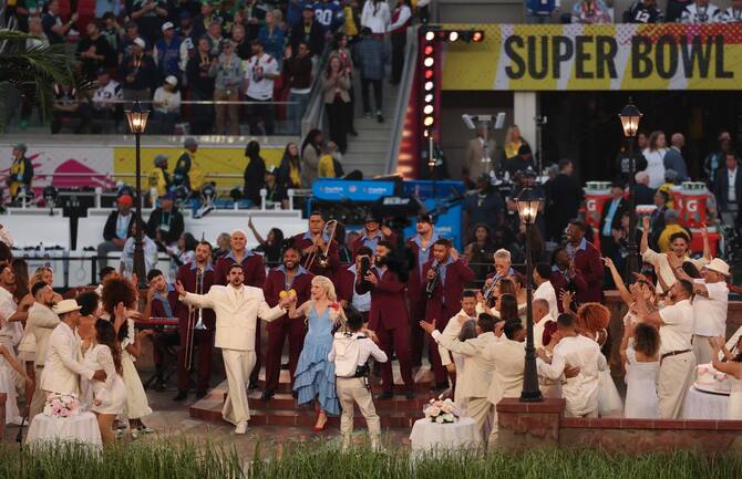 A crowded outdoor stage performance during a Super Bowl event featuring singers in white and blue outfits accompanied by a brass band in maroon suits.