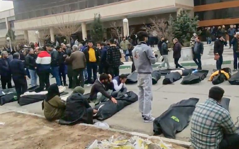 A group of people gathered in an outdoor courtyard where several black body bags are laid out on the ground.