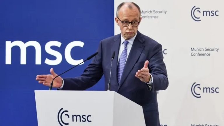 Friedrich Merz speaking at a podium during the Munich Security Conference (MSC), wearing a blue suit and glasses, with a large blue and white "msc" logo in the background.