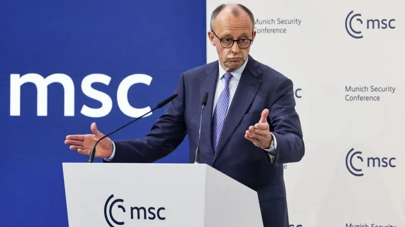 Friedrich Merz speaking at a podium during the Munich Security Conference (MSC), wearing a blue suit and glasses, with a large blue and white "msc" logo in the background.