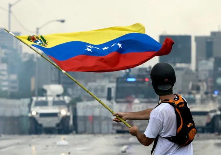 Protester holding a large Venezuelan flag in front of military armored vehicles on a city street.