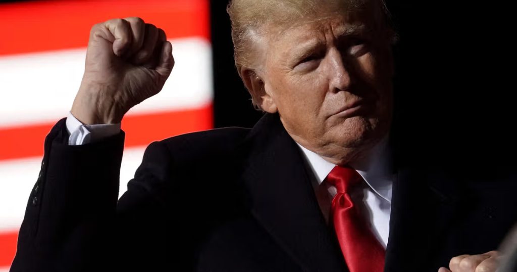 A close-up portrait of Donald Trump raising a clenched fist, wearing a dark coat and a bright red tie, with a blurred American flag featuring red and white stripes in the background.