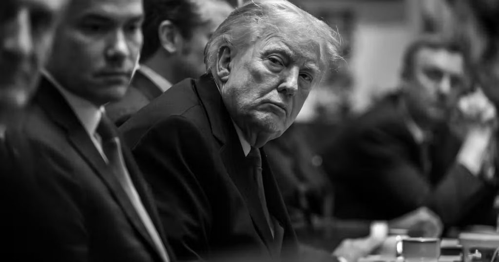 A black and white, high-contrast close-up of Donald Trump sitting at a long table during a meeting, looking towards the camera with a stern expression, surrounded by other men in suits who are partially blurred.
