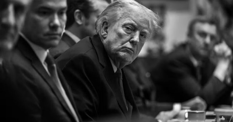 A black and white, high-contrast close-up of Donald Trump sitting at a long table during a meeting, looking towards the camera with a stern expression, surrounded by other men in suits who are partially blurred.