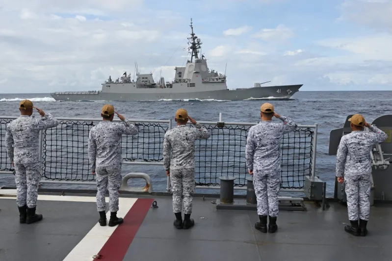 Five sailors in gray and white camouflage uniforms standing on the deck of a ship, facing away from the camera and saluting a large gray guided-missile destroyer with the hull number "41" sailing parallel to them in the open sea.
