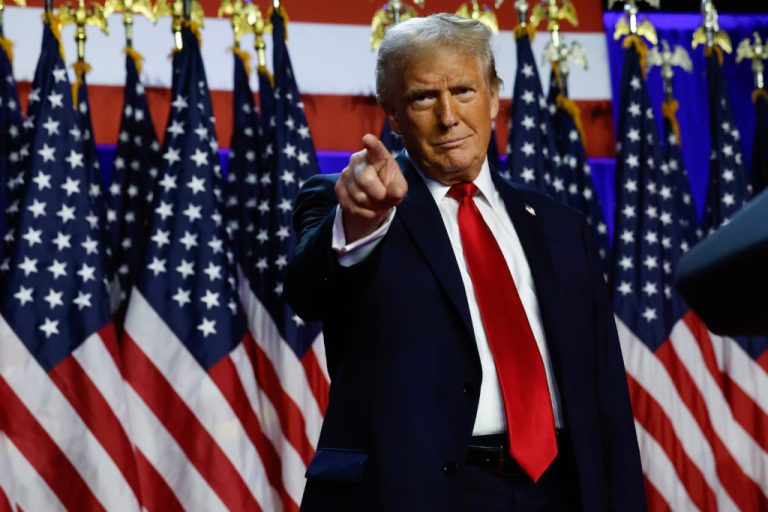 Donald Trump in a dark suit and bright red tie, pointing his finger directly at the camera while standing in front of a row of multiple American flags.
