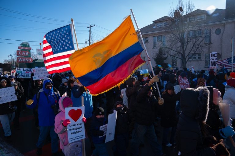 A diverse group of protesters marching on a city street, carrying various flags including the American and Colombian flags, and holding signs such as "Love Your Immigrant Neighbor" with a red heart.
