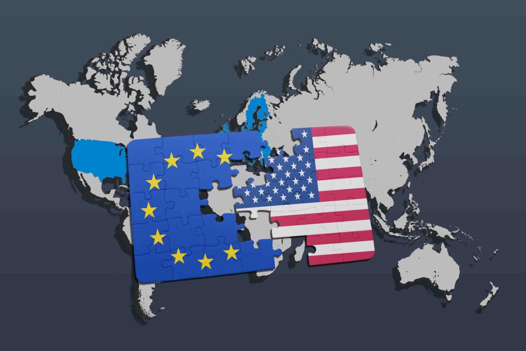 A conceptual digital illustration of a gray world map background with two large puzzle pieces in the center; the left puzzle piece displays the blue flag and yellow stars of the European Union, and the right piece displays the stars and stripes of the United States flag.