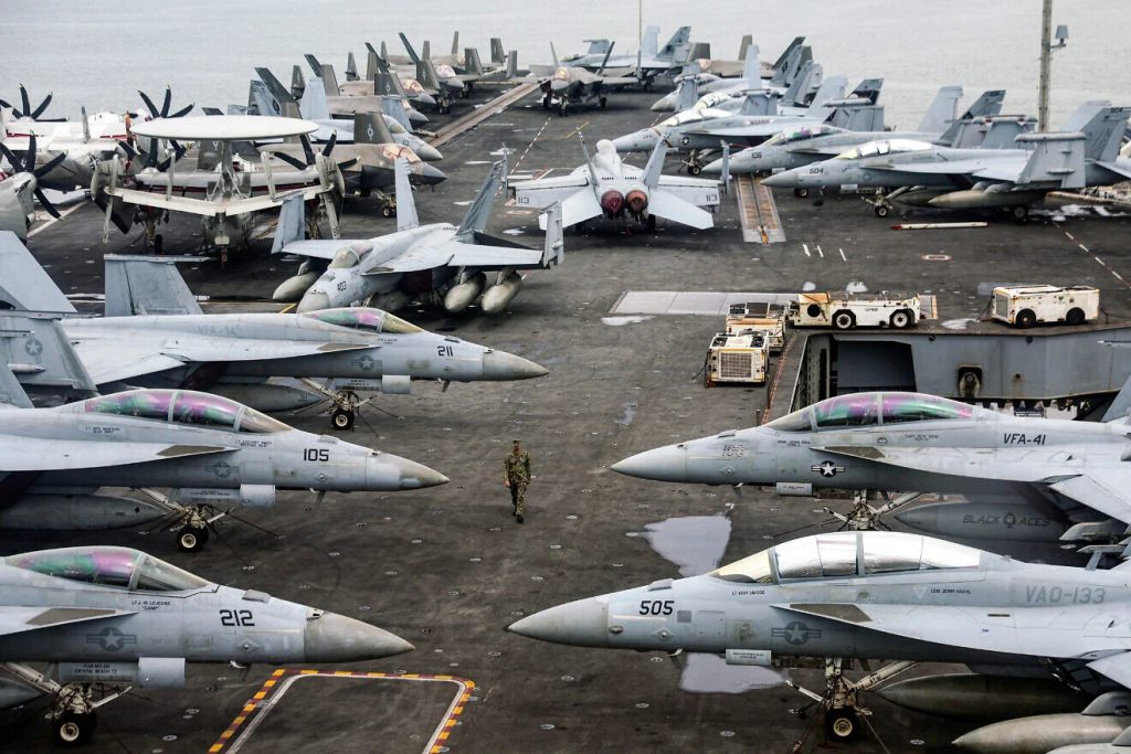 U.S. Navy fighter jets, including F/A-18 Super Hornets and E-2 Hawkeyes, crowded on the flight deck of an aircraft carrier.