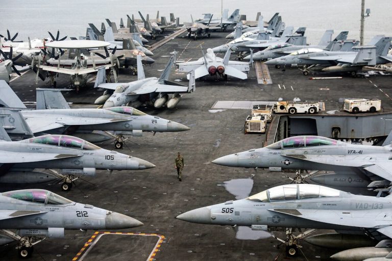U.S. Navy fighter jets, including F/A-18 Super Hornets and E-2 Hawkeyes, crowded on the flight deck of an aircraft carrier.