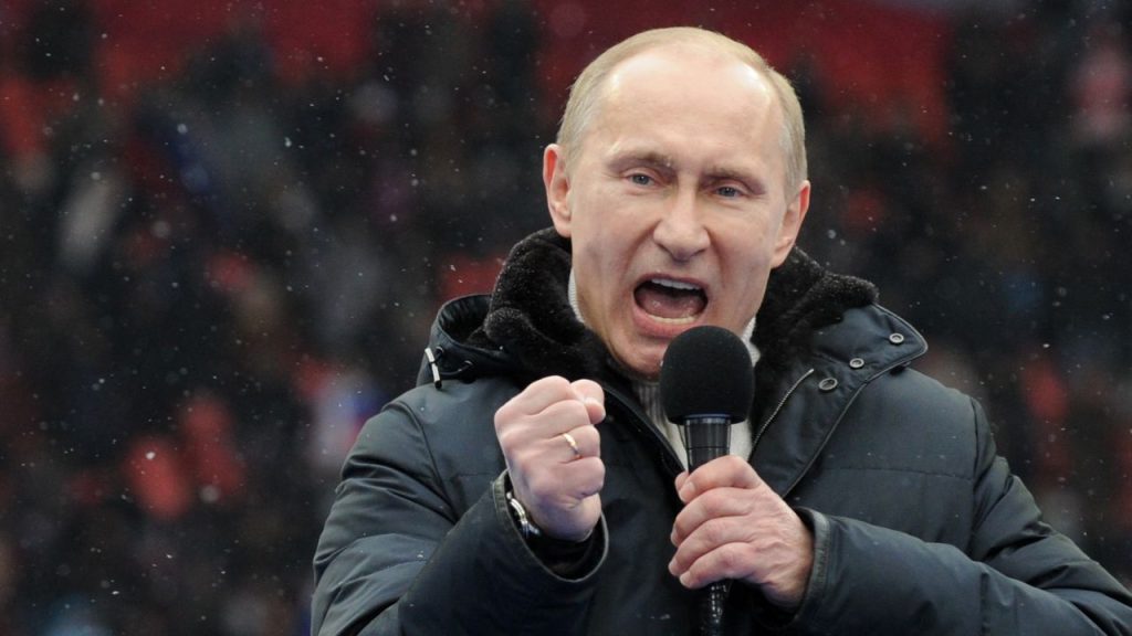 Russian President Vladimir Putin speaking into a microphone with his fist clenched during a large outdoor rally.