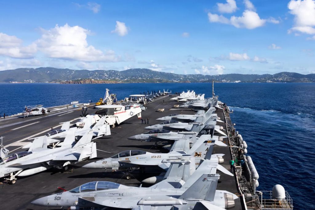 A high-angle view of a United States aircraft carrier deck at sea, packed with various fighter jets including F/A-18 Super Hornets, with a mountainous coastline and blue sky in the background.