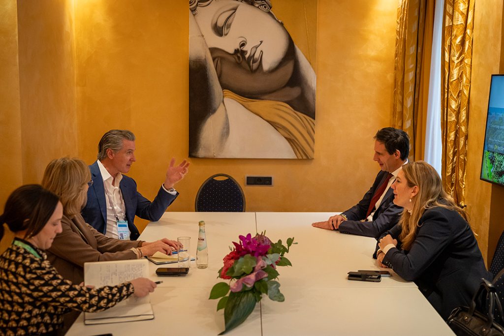 Gavin Newsom in a blue suit sitting at a white conference table, gesturing while speaking to three other people in a room with warm yellow walls and a large artistic painting of a face.