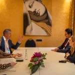 Gavin Newsom in a blue suit sitting at a white conference table, gesturing while speaking to three other people in a room with warm yellow walls and a large artistic painting of a face.