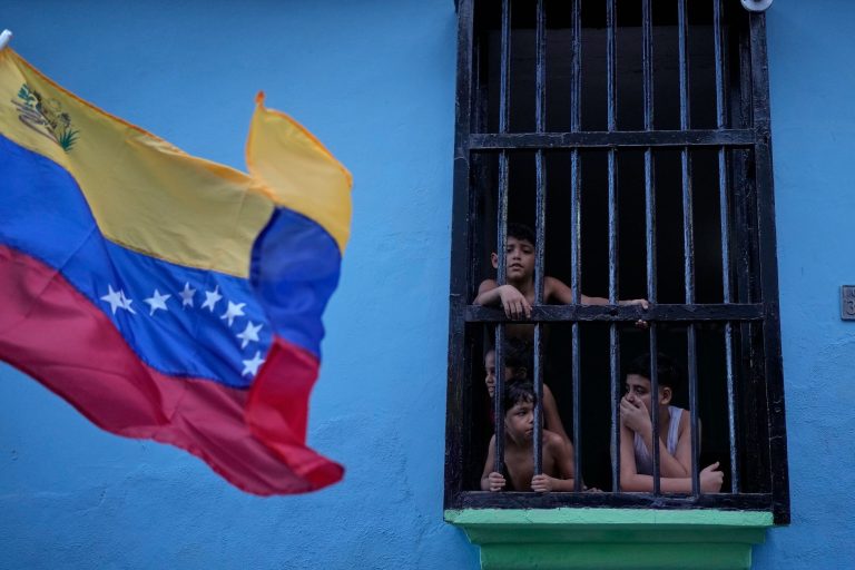 A group of young boys looking out from behind a black iron-barred window of a bright blue building, with a blurred Venezuelan flag waving in the foreground.