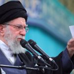 A man with a white beard, wearing a black turban and glasses, speaking into two microphones and pointing his finger.