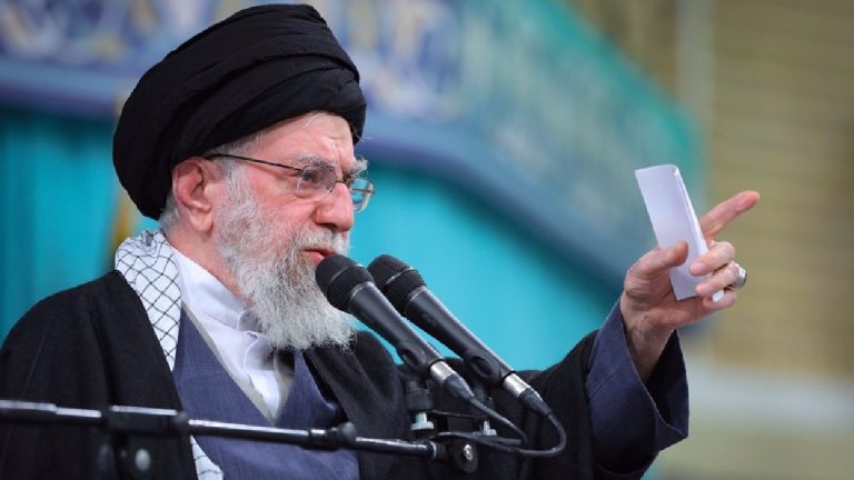 A man with a white beard, wearing a black turban and glasses, speaking into two microphones and pointing his finger.