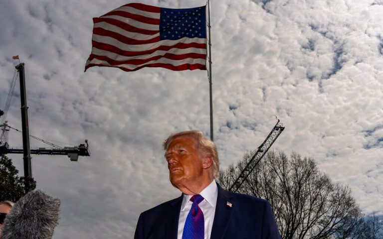 Donald Trump standing under a large waving American flag with construction cranes in the background.