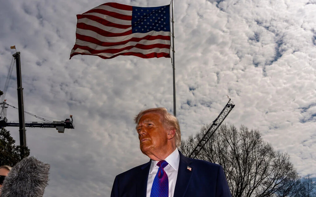 Donald Trump standing under a large waving American flag with construction cranes in the background.