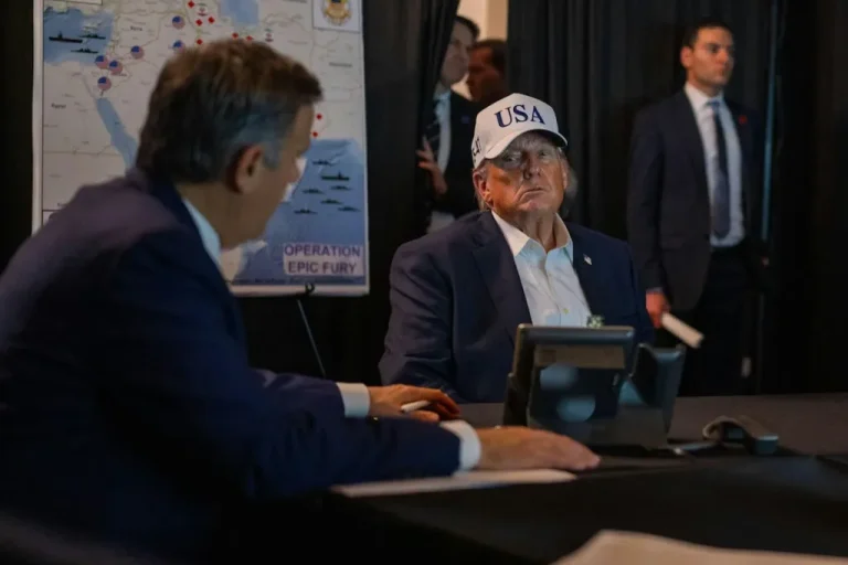 Donald Trump wearing a USA hat sitting at a briefing table with a military map labeled Operation Epic Fury in the background.