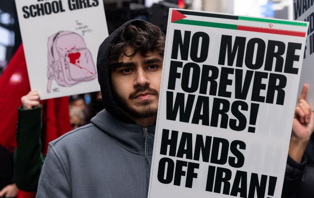 A young man with a beard wearing a dark grey hooded sweatshirt holds a white protest sign with bold black text that reads "NO MORE FOREVER WARS! HANDS OFF IRAN!". In the background, another sign shows a drawing of a pink backpack with red stains.