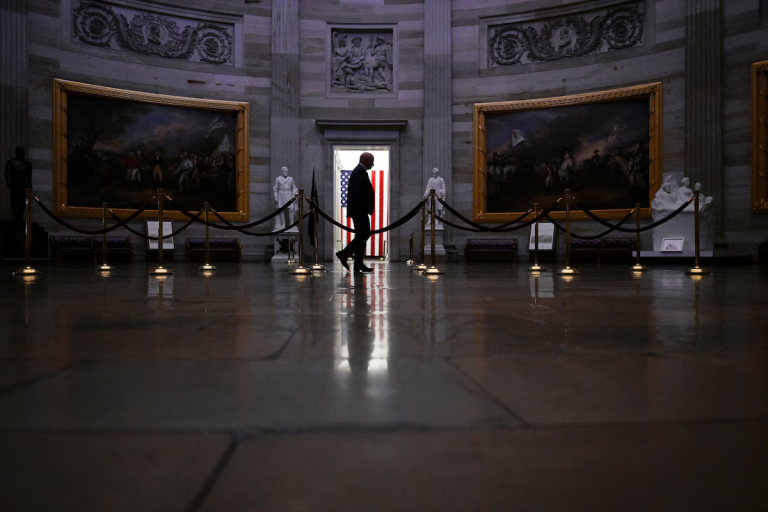 Joe Biden silhouetted walking in US Capitol Rotunda towards a backlit American flag.