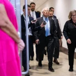 A protester in a helmet and backpack holding a large Venezuelan flag in a city street with armored vehicles in the background.