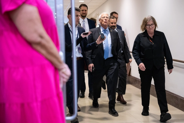 A protester in a helmet and backpack holding a large Venezuelan flag in a city street with armored vehicles in the background.