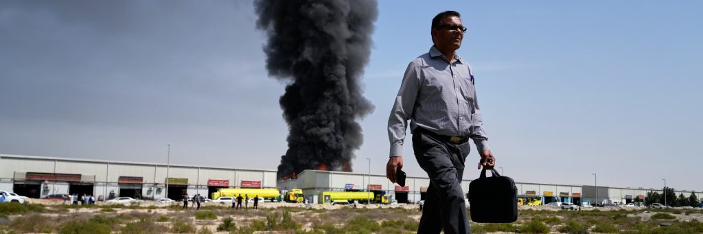 Man walking with a briefcase in front of a massive black smoke plume from a burning warehouse.