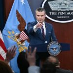 A man in a blue suit and striped tie pointing his finger forward while standing at a podium with "The Pentagon" logo.