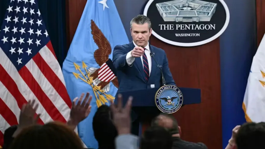 A man in a blue suit and striped tie pointing his finger forward while standing at a podium with "The Pentagon" logo.