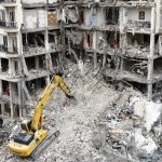 A yellow Komatsu excavator clearing rubble from a heavily damaged multi-story residential building.