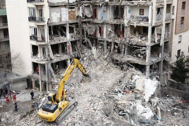A yellow Komatsu excavator clearing rubble from a heavily damaged multi-story residential building.