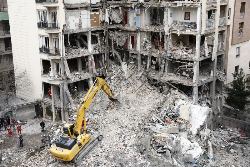 A yellow Komatsu excavator clearing rubble from a heavily damaged multi-story residential building.