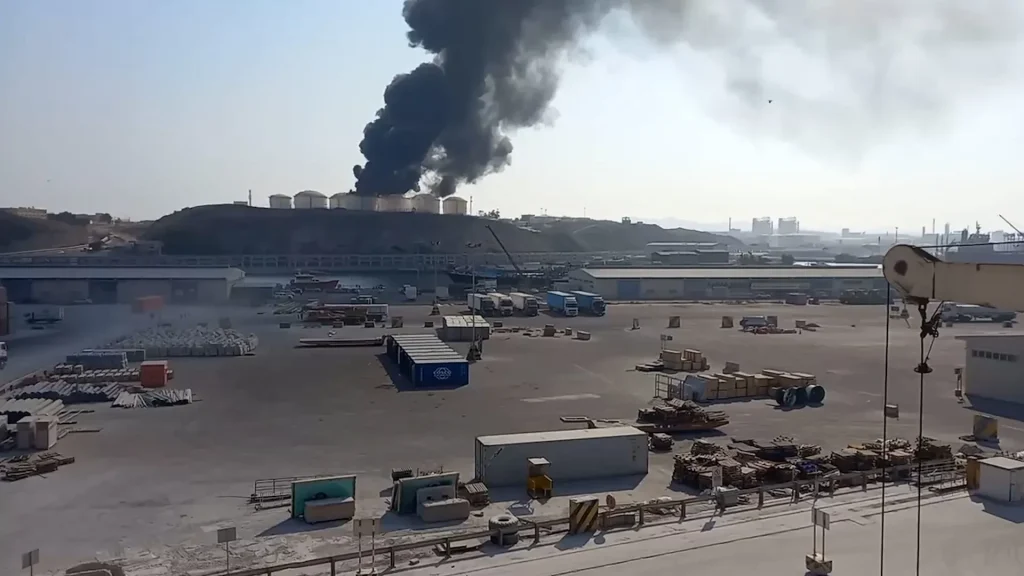 Thick black smoke rising from industrial oil storage tanks near a port facility.