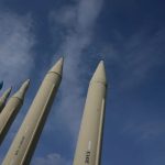 A line of vertically positioned large rockets and iranian ballistic missiles under a blue cloudy sky.