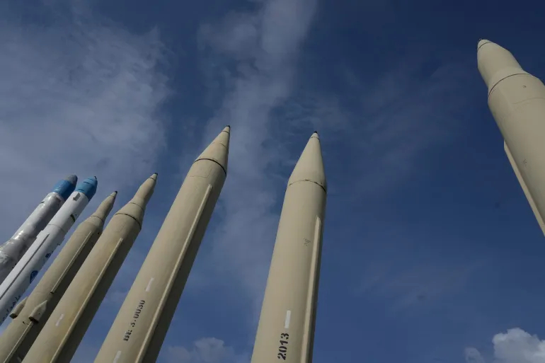 A line of vertically positioned large rockets and iranian ballistic missiles under a blue cloudy sky.