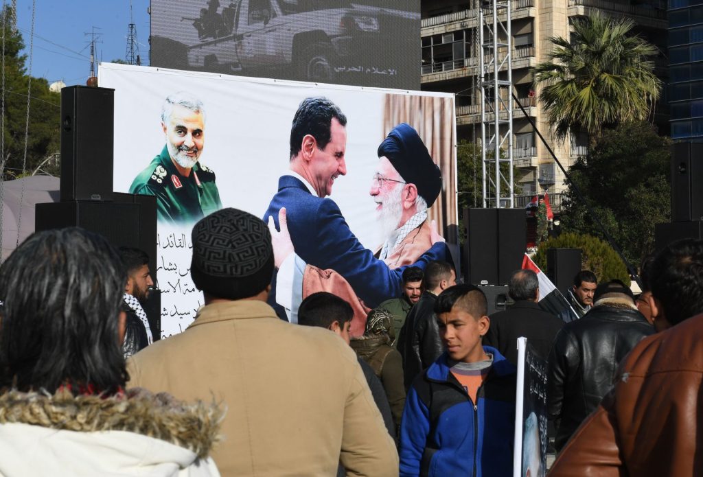 A crowd of people gathered in front of a large outdoor poster featuring three prominent men in a public square.