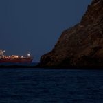 A large red cargo ship sailing near a dark, rocky cliff at twilight or night.