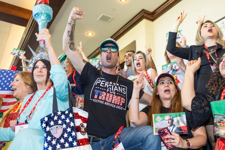 A crowd of supporters at a rally, with a man in a "Persians for Trump" t-shirt shouting and raising his fist.