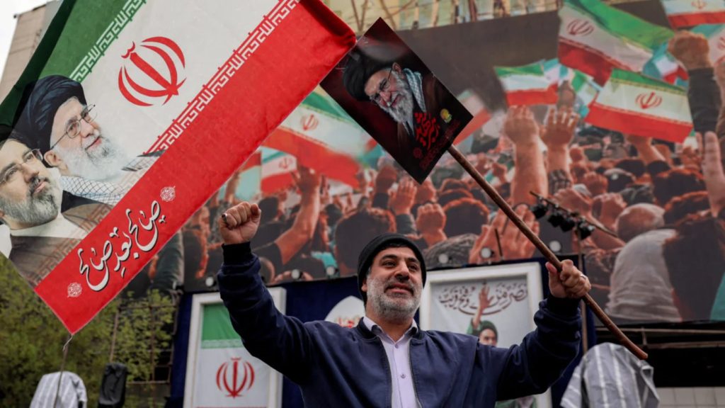 Bearded man holding an Iranian flag and portraits of Supreme Leaders in a large pro-government rally, including a poster of Ayatollah Khamenei.