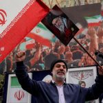Bearded man holding an Iranian flag and portraits of Supreme Leaders in a large pro-government rally, including a poster of Ayatollah Khamenei.