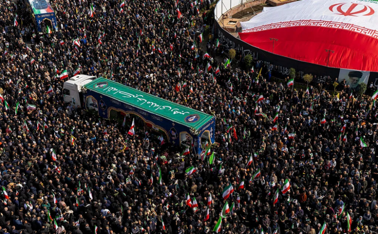 A high-angle, wide shot of a massive crowd of people gathered in an outdoor urban space. A truck decorated with green banners and portraits is moving through the crowd. A large Iranian flag is partially visible on the right side.