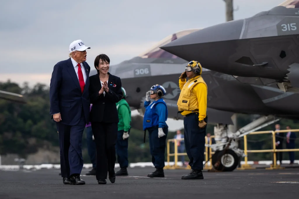 Donald Trump and Sanae Takaichi walking together on an aircraft carrier deck next to a military fighter jet.