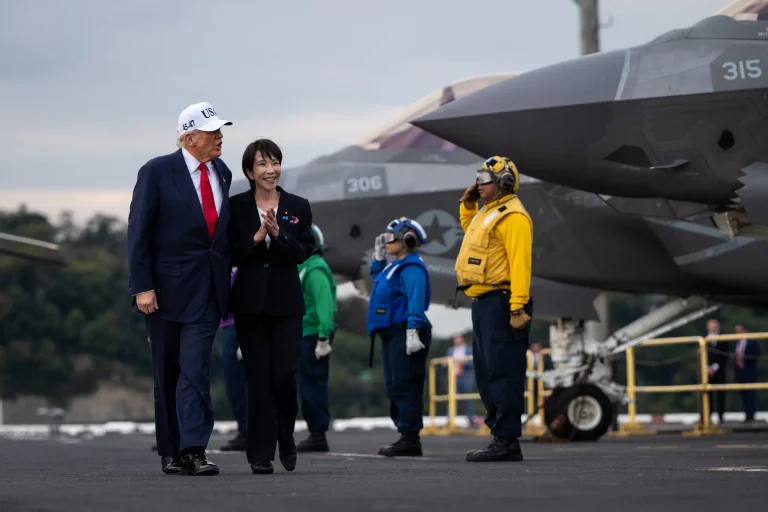 Donald Trump and Sanae Takaichi walking together on an aircraft carrier deck next to a military fighter jet.