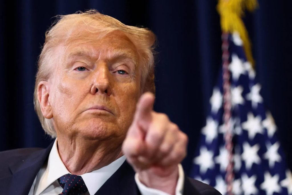 A close-up photograph of Donald Trump looking directly at the camera and pointing his index finger forward, with an out-of-focus American flag in the background.