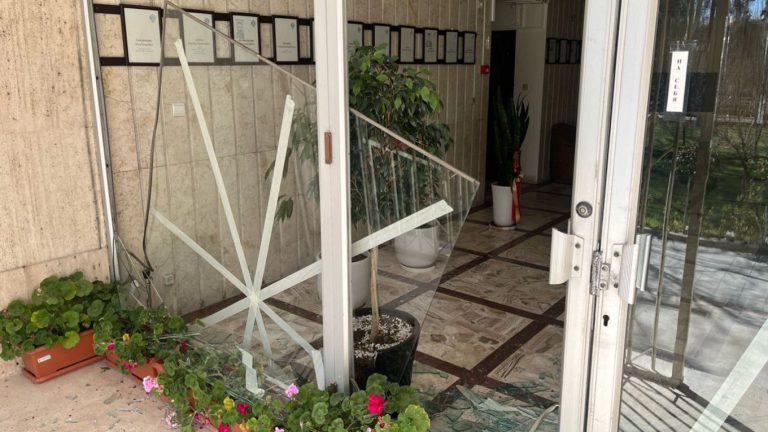 Shattered glass entrance of a government building with broken windows and debris on the floor.