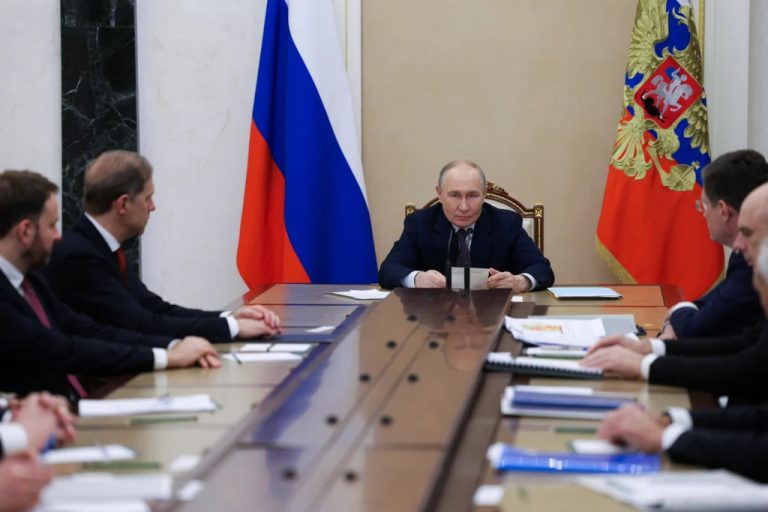 Vladimir Putin presiding over a high-level government meeting at a long wooden table with Russian flags in the background.