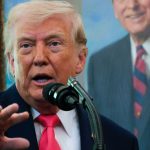 Donald Trump speaking at a microphone with a portrait of Ronald Reagan in the background.